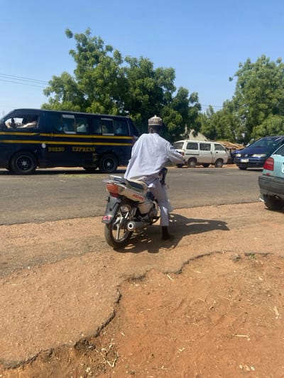 The scene depicts a street in Konkiyel, Nigeria, under a clear, light blue sky, indicating daylight conditions. In the immediate foreground, the ground consists of eroded, reddish-brown soil and degraded asphalt, featuring visible potholes and uneven surfaces. A male individual is positioned on a light-colored Jinchen brand motorcycle, observed from the rear. He wears a light-colored, long-sleeved garment, light-colored trousers, and a patterned cap (kufi). His feet are near the ground, and the motorcycle casts a distinct shadow to its right. The motorcycle itself displays "JINCHEN" on its side and "G.T." on its rear fender.
Adjacent to the foreground, on a paved asphalt road, a dark-colored passenger van labeled "BORNO EXPRESS" in yellow lettering is in motion, traveling from left to right. Multiple occupants are discernible within the van through its windows. Further down the road, to the right of the "BORNO EXPRESS" van, other vehicles are visible, including a white van and a blue sedan, with a partial view of another light-colored vehicle. The background is dominated by dense green foliage from trees, behind which a light-colored structure is partially visible.