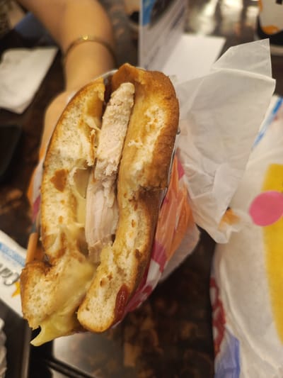 A close-up, slightly tilted shot focuses on a toasted sandwich, cut in half and held by a person. The sandwich features golden-brown bread, revealing generous slices of cooked meat, likely turkey or chicken, along with gooey melted cheese that visibly oozes from the bottom. A hint of red sauce is also visible on the interior of the bread.

The sandwich is partially encased in white paper or plastic packaging. In the blurred background, a person's arm is visible, adorned with a gold bracelet or ring. The setting appears to be indoors, with a dark table surface in the foreground and a mix of white paper and colorful packaging (featuring yellow, pink, and blue/white patterns) in the background. Partial text reading "Car" is visible on a white object in the lower-left. The image depicts a casual meal, possibly at a cafe or restaurant, and based on the provided context, it could be taking place in Samborondón, Ecuador.