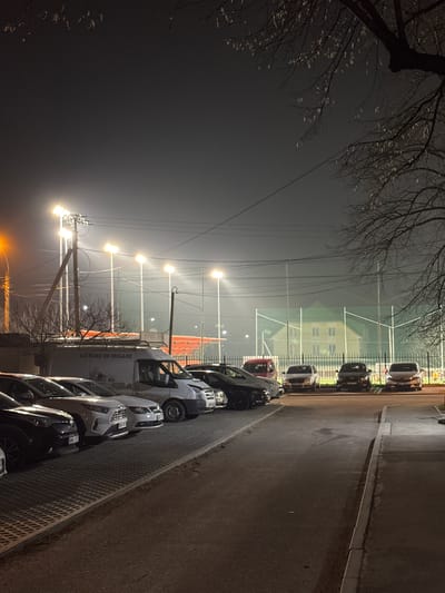 The image depicts a street scene at night in Ciorescu, Moldova. The street is lined with parked cars. A tall fence with lighting behind it runs parallel to the street. Behind the fence, a building is visible. Bare trees are present on the left side of the frame. The sky is dark and overcast. The street is wet.
