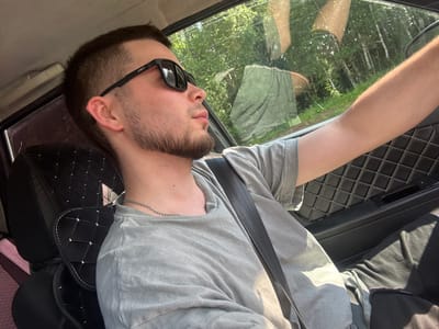 A young man with short brown hair and a beard is sitting in the driver's seat of a car, looking out the window. He is wearing sunglasses, a grey t-shirt, and a silver chain. The car's interior is visible, including a black seat with diamond stitching and a seatbelt. Outside the window, there is a reflection of trees and sky, indicating the car is in a natural setting, possibly a forest or rural area. The image appears to be a selfie taken by the driver.

The location is Nytva, Russia. This is implied by the user's prompt requesting the location in Nytva, Russia. Without additional context or visual cues specific to Nytva within the image itself, it is impossible to definitively confirm the location. However, the surrounding greenery and trees are consistent with a Russian landscape.
