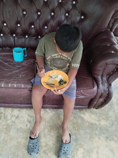 A child sits on a worn, dark brown tufted sofa adorned with silver-colored buttons, holding an orange circular plate containing white rice, dark green leafy vegetables, and a piece of protein covered in a brown sauce. A silver-colored spoon is positioned in the child's right hand over the plate. To the child's left, on the sofa cushion, rests a turquoise-blue plastic mug with a handle. The child is wearing an olive-green t-shirt with faded text and design, and blue shorts. The child's bare feet are partially inserted into dark grey, perforated clog-style shoes. The floor beneath is a light-colored concrete surface, exhibiting mottled grey and tan patterns. This scene is located in Teupinbayu, Indonesia.