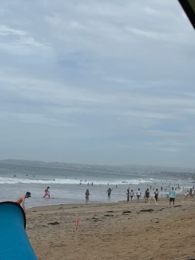 The image depicts a crowded beach scene on a cloudy day. In the foreground, a blue tent is partially visible on the sandy beach. A person wearing a blue baseball cap and red shorts is standing near the tent. A red shovel is stuck in the sand to the right of the tent. The middle ground shows the ocean with many people swimming and playing in the waves. A distant coastline with buildings is visible in the background. The overall atmosphere suggests a typical summer day at a popular beach. Based on the visual cues of the coastline and the type of beach, this scene is likely located in San Clemente, California, United States of America.