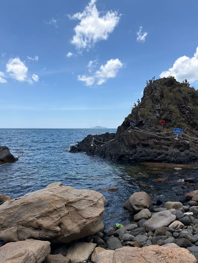 The image depicts a rocky shoreline and a dark volcanic rock formation jutting out into the ocean under a bright blue sky with scattered white clouds. The water is a deep blue with ripples. Several large, light-brown boulders are in the foreground, with smaller dark rocks and pebbles leading down to the water. On the volcanic rock formation, there are signs and a rope barrier, suggesting it is a designated area or a point of interest. Sparse green vegetation, possibly cacti, grows on the top of the formation. The scene is located in Phú Lương, Vietnam, and appears to be a natural landscape with some minor human intervention for visitor management. There are no people visible in the image.