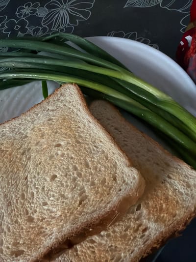 The image shows two slices of toasted bread on a white plate, next to a bunch of green onions. The bread appears to be whole wheat, with a slightly browned crust and a porous, light brown interior. The green onions are fresh and have a vibrant green color, with some visible moisture on their surface. The plate is round and white, with a slightly raised edge. The background is a dark surface with a floral pattern in white. A glimpse of red, possibly from food packaging, is visible in the upper left corner. The scene is shot from a high angle, looking down at the food. There is no visible text in the image. The lighting suggests an indoor setting, likely during the day. The overall impression is of a simple, fresh meal or ingredients prepared for a meal.