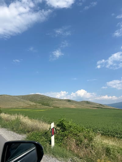This image depicts a rural landscape in the vicinity of Isperihovo, Bulgaria, under a clear blue sky adorned with scattered white cumulus clouds. In the background, undulating hills covered in dry, light green, and brown vegetation stretch across the horizon. A slender, light-colored tower or mast stands atop the prominent hill in the left-center. Below the hills, agricultural fields are visible; a vast expanse of vibrant green, low-lying row crops dominates the lower midground and extends to the right. Patches of yellowish and greener vegetation are present closer to the base of the hills on the left. The foreground features the edge of a road with a grey gravel shoulder, bordered by a dense strip of wild vegetation composed of dry grasses and green bushy plants. A white rectangular reflector post with a vertical red stripe is positioned within this vegetation, slightly to the right of the lower center. The top portion of a vehicle's side mirror is visible in the bottom-left corner, reflecting internal elements of the vehicle.