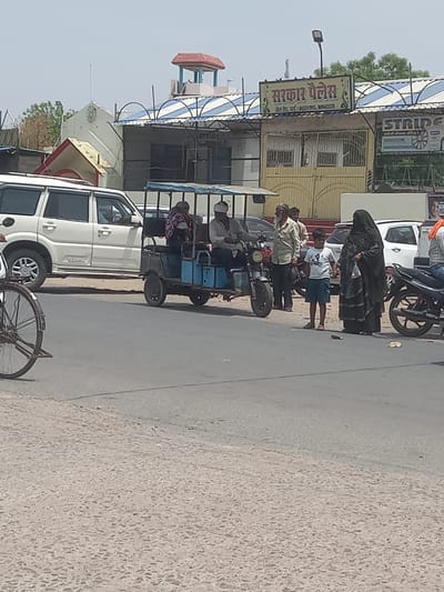 Captured in a medium, full-shot view, the scene unfolds on what appears to be a street in Orai, India, specifically on Jail Road. The day is bright, with the sun illuminating the scene under a clear sky. The street scene is moderately busy, with an assortment of vehicles and pedestrians populating the area. A white SUV, possibly a Scorpio model, is prominently featured near a blue and black auto rickshaw. The rickshaw is carrying several passengers. Notably, an individual is dressed in a black Burqa with a child standing close by. Other pedestrians can be seen conversing or waiting. On the left side of the image, partially obscured by the SUV, sits a building adorned with a sign that reads "Sarkar Palace" and its contact information, indicating it is a commercial establishment. Another sign for "Stride" is partially visible on the right, advertising English speaking classes. The surrounding environment is a mix of buildings, signs, and the road itself, indicative of a typical urban street. The road appears to be made of concrete.