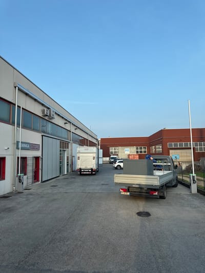 The image depicts an industrial or commercial loading area in Sarcedo, Italy, under a clear blue sky. The scene is devoid of people.
In the foreground, a dark grey flatbed truck, license plate "FS 863EL," is parked facing away from the viewer. It carries a large, grey, rectangular cabinet-like object and other miscellaneous items. A dark oil stain is visible on the asphalt beneath the truck. To the right of the flatbed truck, a white vertical barrier arm pole with a green and grey control unit stands.
On the left, a long, light-colored industrial building features multiple horizontal windows, some with internal blinds, and red-framed doors. The text "BENINCA STORE" is visible on the building's facade. Two white air conditioning units are mounted on the wall above the windows. A white box truck, displaying "norder" on its rear, is backed up towards an open loading bay or entrance on this building. Another white vertical barrier arm pole with a green and grey control unit is positioned near the entrance of the "BENINCA STORE."
In the background, behind the flatbed truck and between the main buildings, several parked vehicles, including a white SUV and a white van, are visible. Further back, a red-brown building with multiple windows features a large "HI" logo on its exterior. Red storage containers and a basketball hoop are also visible in the vicinity of this red-brown building. The ground consists of paved asphalt and concrete, with some green foliage visible along the right edge of the frame, behind a fence.