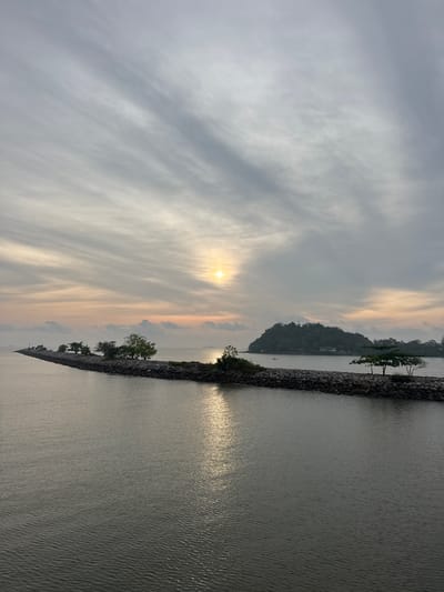 The image shows a sunrise or sunset scene in Pak Nam Chumphon, Thailand. A body of water, likely the sea, occupies the right side of the frame, reflecting the sunlight. A rock barrier with sparse vegetation separates the water from the sky. The sky is filled with wispy clouds, with the sun partially visible. A small island is visible in the distance.
