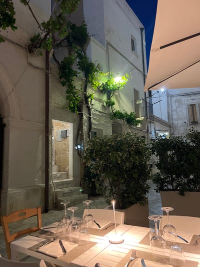 The image shows an outdoor dining setting in Polignano a Mare, Italy. A white-clothed table is set with wine glasses, napkins, and cutlery. A small, modern lamp illuminates the setting. A simple wooden chair sits at the table. The architecture of the surrounding buildings is white, with stone accents. Vines and trees grow on the buildings. Steps are visible with Italian writing on them. An awning is partially visible overhead. It is evening or night, with limited natural light.