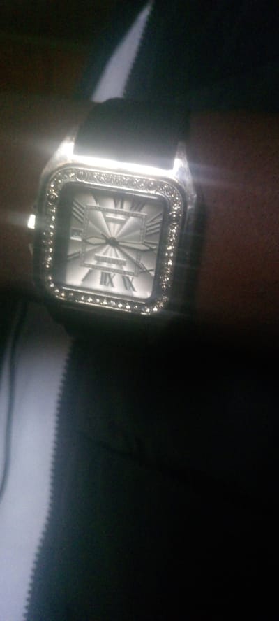 The image shows a close-up of a square wristwatch with a silver-toned case and a white dial. The dial features Roman numerals and a sunburst pattern radiating from the center. The bezel of the watch is decorated with small, clear stones that appear to be rhinestones. The watch is on a dark-colored surface, possibly skin or fabric, and there is a bright light source reflecting off the watch face. The background is dark and out of focus. There is no information in the image to suggest it is located in Zot Shen, Nigeria.