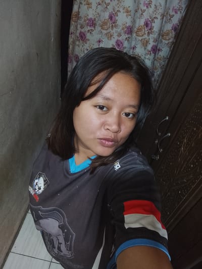 A young woman is photographed indoors, standing in what appears to be a narrow hallway or doorway. She is looking directly at the camera with a neutral expression. She has dark hair, pulled back slightly, and is wearing a dark t-shirt with blue trim around the collar. The t-shirt features a graphic design on the left chest and a red, white, and blue stripe on the right sleeve. To her left is a plain, gray textured wall, and to her right is a wooden cabinet or door with decorative carvings and three handles. Behind her, a floral curtain is visible, with a pattern of roses in pinks and creams. The floor appears to be tiled. The image is a close-up, capturing her from the chest up, with the background blurred. The lighting suggests an indoor environment, possibly dimly lit. The presence of a wooden cabinet and floral curtains could indicate a residential setting. The exact location within Gunung Malang, Indonesia, cannot be determined from the visual information alone.