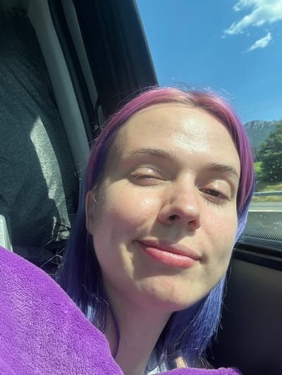The image shows a selfie of a fair-skinned person with purple hair, likely taken inside a vehicle. The person's eyes are partially closed, and they have a slight smile. Part of a window and interior of a vehicle is visible, with a view of a blue sky and possible foliage outside. The location is presumed to be Pourrières, France, although this cannot be confirmed from the image alone. There is a purple, possibly fleece, item of clothing resting on the person's chest.