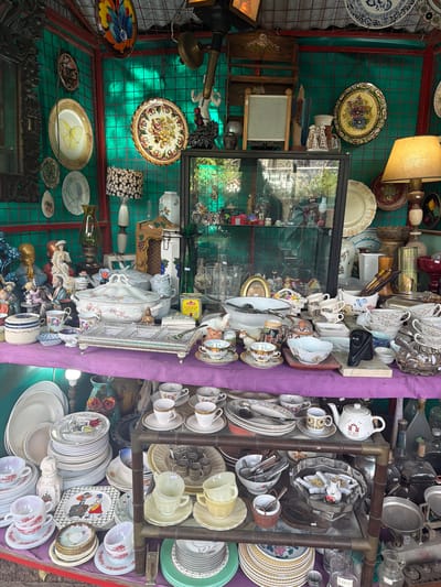 This image depicts a stall at a flea market in Acassuso, Argentina, showcasing a cluttered array of antique and vintage items. Predominantly displayed are porcelain teacups, saucers, plates, and serving dishes. These are organized on shelves and stacked on a purple-draped table. There are lamps, decorative bowls, figurines, and other collectibles on a green wire backdrop. The photo is taken from an angle, with the items positioned as if it were inside the market stall. A glass case in the middle displays more fragile items. The stall is illuminated by natural light.