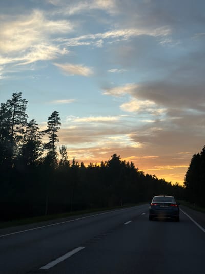 A dark-colored sedan, bearing the license plate NN-8921, is driving down a two-lane asphalt road, heading away from the viewer. The vehicle's taillights are illuminated. The road is lined with trees on either side, silhouetted against a sky filled with wispy clouds. The sky exhibits a gradient of colors, transitioning from light blue at the top to hues of orange and yellow near the horizon, indicating either sunrise or sunset. The environment appears to be rural, possibly within the vicinity of Usma, Latvia, given the presence of dense forestation. The lighting suggests it is either early morning or late evening.
