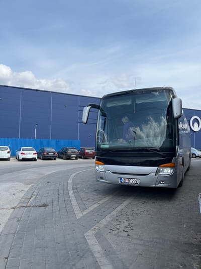 The scene depicts a silver-grey Setra touring coach, bearing license plate BD BE 292, parked on a grey, brick-paved area at the BIG Fashion shopping center in Cacak, Serbia. A single individual, dressed in a light-colored top, is partially visible within the bus's front cabin through the windshield. The background features a large, rectangular commercial building with a dark blue corrugated metal facade, displaying the white circular logo of 'BIG Fashion Cacak' on its right side. A row of various parked passenger vehicles, including a white BMW sedan, a dark Mercedes-Benz sedan, and a red compact car, is situated further back against the building. White directional markings are painted on the paved ground. The sky is partly cloudy, with cloud reflections visible in the bus's windshield.