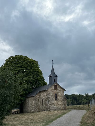 The image shows a small stone chapel with a dark slate roof and a steeple topped with a weather vane. The chapel is situated in a grassy field, with a large, leafy tree to its left and a gravel path leading away from it towards the right. In the background, a line of trees forms the edge of a forest, and a fence runs along the right side of the frame. The sky is overcast with heavy, gray clouds, suggesting a moody or impending weather event. The chapel appears to be located in a rural or semi-rural setting, possibly in or near the village of Bech, Luxembourg, given the context. There are no people visible in the image. A small dark object, possibly a bench or a seating area, is visible under the large tree.