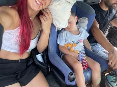 A mother with bright pink hair, wearing a white tank top and black shorts, smiles while taking a selfie in a car. Next to her, a young boy sleeps in a car seat, leaning against a pillow. A man in a black t-shirt, presumably the driver, is visible in the background. A bottle of red liquid and a black bag are also present in the car. The scene is set on a sunny day, likely in Orliak, Bulgaria, as indicated by external context.