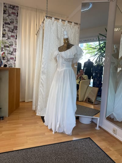 In a well-lit room, likely a bridal shop or tailor's studio in Oldenburg, Germany, a white wedding dress is displayed on a mannequin. The dress features a ruffled neckline and a full skirt, with a shimmering gold sequined bustier underneath. Behind the dress, a cream-colored curtain with a diamond pattern hangs, partially obscuring a beige chair. A man, appearing to be a photographer, is captured in the reflection of a mirror hanging on the wall, taking a picture of the dress with his phone. He is wearing a light-colored t-shirt and dark pants. The room has wooden flooring and a dark patterned rug. In the background, a plant is visible, and reflections in the mirror show mannequins in patterned clothing, hinting at a retail or fitting environment. The scene suggests a moment of preparation or showcase, possibly for a wedding or a photography session.