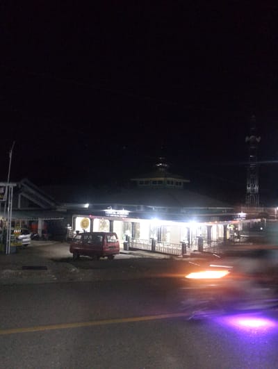 The image depicts a nighttime scene in Pompanua, Indonesia. The primary subject is a mosque, identified by the visible text "MASJID AL-MUQARRAMEN" and "JEMBATAN KEL. PERPUA." The mosque has a distinctive green, multi-tiered roof and is illuminated by numerous lights, creating a warm glow. In front of the mosque, a white railing separates the building from a paved area. A red minivan is parked on the paved area, and to its left, a silver vehicle with "TAXI" written on it is also visible. A tall communication tower stands to the right of the mosque, silhouetted against the dark sky. The foreground shows a road with a yellow line, and streaks of orange and purple light suggest the movement of vehicles or possibly light reflections. The overall atmosphere is tranquil, with the artificial lights of the mosque and tower standing out against the dark night. There are no people clearly visible in the image, and the weather appears to be clear.