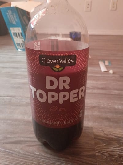 A nearly empty 2-liter bottle of "Dr. Topper" soda, a Clover Valley brand, lies horizontally on a light-colored wooden floor. The bottle is positioned so that the label is facing the viewer. Part of a blue box is visible in the lower left corner of the image. The background also includes a white wall with electrical outlets. The setting is indoors, likely in a home, but there is no indication of a location in Clarksville, United States of America.