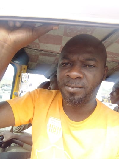 A male individual with dark skin, a shaved head, and a short, dark beard is positioned centrally in the foreground, looking directly at the camera. He wears a bright orange short-sleeved t-shirt with a light-colored emblem on the chest. His right hand is raised, gripping an overhead frame or support within the vehicle.
The scene is set inside an open-sided commercial vehicle, likely a tricycle, in Gyadi Gyadi Arewa, Nigeria. The vehicle's interior features a light-colored roof lining with a red and white checkered pattern. A yellow structural frame is visible on the left side of the vehicle's opening, incorporating a circular reflective object.
Two other individuals are partially visible: one behind and to the left of the central figure, with only the top of their head and shoulder visible, wearing a light-colored garment; and another on the far right, seen in profile, wearing a cap. An arm and hand belonging to a third individual are visible in the lower-left quadrant, near what appears to be a vehicle control panel.
Through the open side of the vehicle, a bright daytime outdoor environment is visible. This includes a paved surface, a light-colored car in the mid-ground, and faint distant structures or hills under a bright sky.