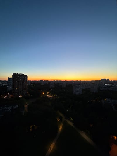 The image depicts a dusky cityscape, likely taken from an elevated vantage point, showing a gradient of sky colors from deep blue at the top to a bright orange band near the horizon. Beneath the sky, a dense urban landscape unfolds, characterized by numerous multi-story buildings of varying architectural styles. In the foreground, a large, dark-colored residential building with illuminated windows is prominent. The mid-ground features a dark expanse of trees, interspersed with streetlights that cast pools of illumination on pathways and roads. The city stretches into the distance, with more buildings visible against the fading light. The overall scene suggests an evening or early morning setting in a densely populated urban area, possibly Boroduki, Russia, given the context provided. No people are clearly identifiable in the image.