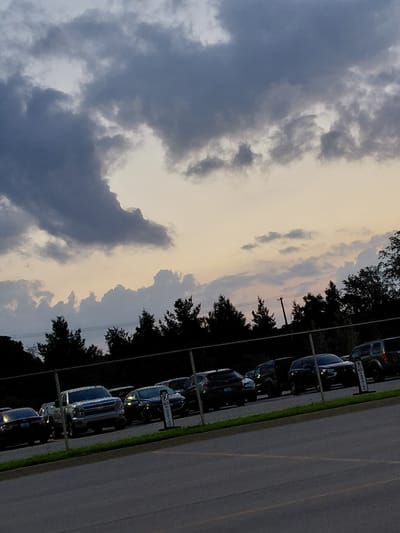 The image captures a parking lot in Lexington, USA, during what appears to be dusk. A chain-link fence runs horizontally across the mid-section of the frame, separating the parking lot from a line of trees and a dusky sky. Several vehicles, including a silver pickup truck, a dark SUV, and a black sedan, are parked in rows behind the fence. The sky is filled with dramatic, dark clouds that are beginning to dissipate, allowing a soft, yellowish light to peek through. The overall impression is one of a peaceful, end-of-day scene.