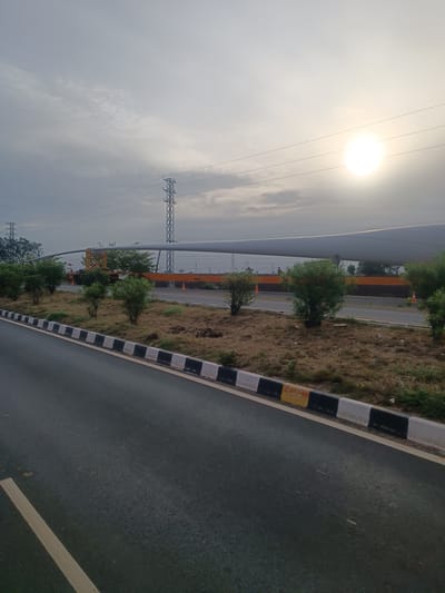 The image shows a view of a road with a large wind turbine blade being transported on a flatbed truck. The scene is set in Thammarajupalle, India, under a hazy, overcast sky with the sun partially visible. The road is asphalt with a white line marking its edge. A curb with alternating black and white painted sections runs along the side of the road, with some sections painted yellow. Beyond the curb, there is dry, brown vegetation and several small green trees. In the background, a large wind turbine blade is mounted on a trailer, supported by a yellow crane or transport vehicle. Several power transmission towers with wires are visible in the distance, along with more trees and vegetation. The overall impression is one of an industrial or infrastructure development site, possibly on the outskirts of a town or rural area. There are no people visible in the image.
