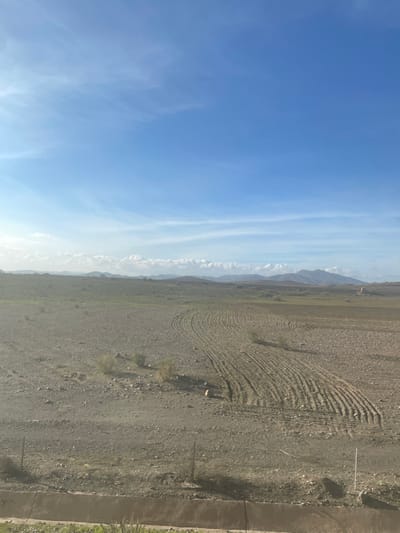 This media file captures a wide-angle landscape scene, likely oriented horizontally despite its current vertical presentation. The upper portion of the corrected image features a vast, clear blue sky, sparsely adorned with soft, wispy white clouds, indicating fair weather. Along the distant horizon, a range of mountains appears hazy, stretching across the frame.
The dominant feature of the lower two-thirds of the image is an expansive, arid field or plain. The ground is a mix of light brown and gray, characterized by rocky and sandy soil. Distinctive parallel furrows, arranged in a large, sweeping curve, are visible across the mid-ground, suggesting the land has been recently tilled or prepared for cultivation. Sparse, dry vegetation, appearing as small clumps of bushes or grasses, is scattered throughout the field.
In the foreground, running along the right side of the image, is a concrete structure, possibly an irrigation canal or a low boundary wall. Several thin, upright poles or stakes are also visible near this structure.
No people or identifiable man-made objects beyond the tilled land and concrete structure are visible. The scene appears static, with no active event taking place, though the furrows imply recent agricultural activity or preparation. The bright, even lighting suggests it is daytime, likely morning or early afternoon. The overall environment points to an arid or semi-arid region, consistent with the location context of Jbilate, Morocco. There is no visible text in the image.