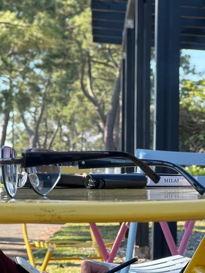 The image depicts a close-up view of sunglasses resting on a reflective surface, likely a table, in an outdoor setting. The sunglasses are black with clear lenses and are positioned in the foreground, angled to the left. Behind the sunglasses, a black cylindrical object, possibly a lighter or a pen case, is visible. Further back, a package or box with the word "MILAN" printed on it is also present. The surface they rest upon reflects the objects and the background. The background consists of blurred trees and foliage, suggesting a park or natural environment. The lighting indicates it is daytime, with shadows visible. The overall scene has a casual, relaxed feel. There is no clear indication of the city of Ochamchire, Abkhazia in the image.