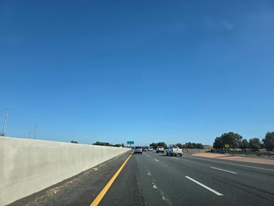 The image depicts a highway scene on a clear, sunny day. The perspective is from a vehicle traveling on the right side of the road, suggesting a location in the United States. The sky is a vibrant, cloudless blue, indicating fair weather and likely mid-morning or afternoon.
The road itself is paved asphalt with distinct white lane markings and a solid yellow line delineating the edge. Several vehicles are visible on the highway, including cars and pickup trucks, all traveling in the same direction. The activity is simply highway driving.
To the left of the highway, a tall, light-colored concrete barrier runs along the side. Beyond the barrier, utility poles with power lines are visible against the sky, and some sparse vegetation. To the right of the highway, there are trees, bushes, and some signs. One sign, with a diamond shape and black symbol, appears to be a warning sign. Further in the distance, a green highway sign is visible, partially obscured, but seems to indicate directions and possibly distances to towns or exits. The presence of a concrete barrier and highway signs points to an established roadway infrastructure.
The overall impression is one of a typical drive on a well-trafficked highway in a rural or semi-rural setting. The lighting suggests the sun is high, casting minimal shadows. No people are clearly discernible within the vehicles. The scene is mundane and functional, focused on transportation.