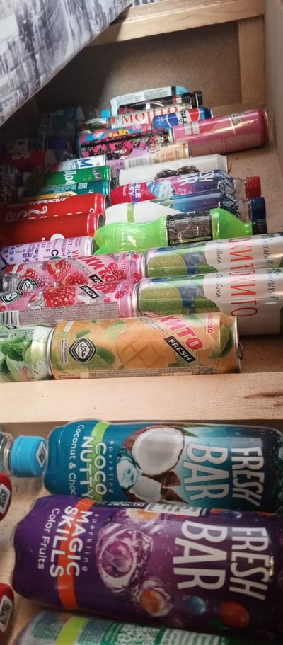 This media file shows a close-up view of a collection of canned and bottled beverages stacked inside a drawer or storage space. The scene appears to be indoors, with a wooden shelf visible in the foreground. The lighting suggests it might be daytime, but the enclosed space makes it difficult to determine the exact time of day or weather conditions.
The subjects are various types of drinks, including what appear to be soft drinks, energy drinks, or flavored water. Several cans and bottles display colorful labels with text in both English and Cyrillic script. Some visible text includes "MOJITO," "FRESH BAR," "COCO NUTTY," "MAGIC SKILLS," and "HALAL." The cans and bottles are arranged in multiple layers, with some partially obscured by others. The overall impression is one of a stored collection of beverages, possibly for personal consumption or retail. There are no people visible in the image. The location context is Khusainovo, Russia.