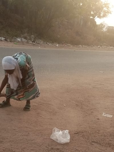 The image captures a woman bending over on the dirt shoulder of an asphalt road in what appears to be a rural or semi-urban setting, identified as Ebiya, Nigeria. She is wearing a light-colored headscarf, possibly white, and a long, patterned dress with shades of light blue, green, and brown. Her feet are clad in dark sandals.
Her posture suggests she is engaged in an activity close to the ground, possibly picking something up or tending to something on the dirt. In the immediate foreground, a crumpled, translucent plastic bag lies on the reddish-brown earth, and other smaller pieces of debris are scattered nearby.
The background shows sparse vegetation, including trees and bushes, on a slightly elevated or hilly terrain. Along the edge of the road and nestled within the background foliage, a considerable amount of litter and discarded materials are visible, indicating an issue with waste accumulation.
The lighting is soft and hazy, with a warm glow that suggests either early morning or late afternoon sun, diffused and creating a slight glare in the upper part of the image. The general atmosphere is somewhat dusty. There is no visible text in the image, and the woman's face is obscured, preventing any clear emotional cues.