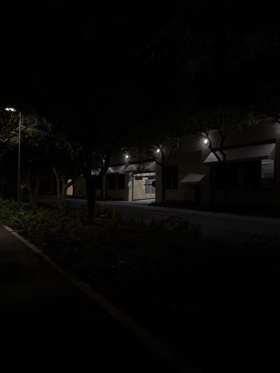 A dimly lit alleyway at night features a multi-story building on the left with glowing lights along its façade. Trees with sparse foliage line the alleyway, their branches reaching towards the building. On the right is a dark wall or barrier, obscuring most of the scene. The composition is shot from a low angle, tilted slightly to the left. The overall atmosphere is quiet and mysterious.