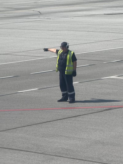 A male airport ground crew worker, wearing a dark uniform, a bright yellow safety vest, and headphones, stands on a paved surface marked with white and red lines. He is facing left, with his right arm extended horizontally, palm down, in a signaling gesture. The scene appears to be an airport tarmac under a bright, sunny sky. The background consists of the same paved surface, showing various painted lines and subtle surface variations. The location is specified as the city of Kalinino, Russia.