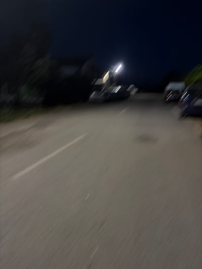 The image depicts a blurry night scene, suggesting motion. A paved road with a white lane marker runs diagonally across the frame. In the background, several vehicles are parked or stopped on the side of the road. One vehicle appears to be a light-colored van or truck, while others are darker sedans or SUVs. A street light is visible in the middle ground, emitting a bright glow. The sky is dark, indicating it is nighttime. The overall impression is of a quiet, possibly residential or roadside area in Kukorevo, Bulgaria, captured during the evening or night. There are no people clearly visible in the image due to the blur and darkness.
