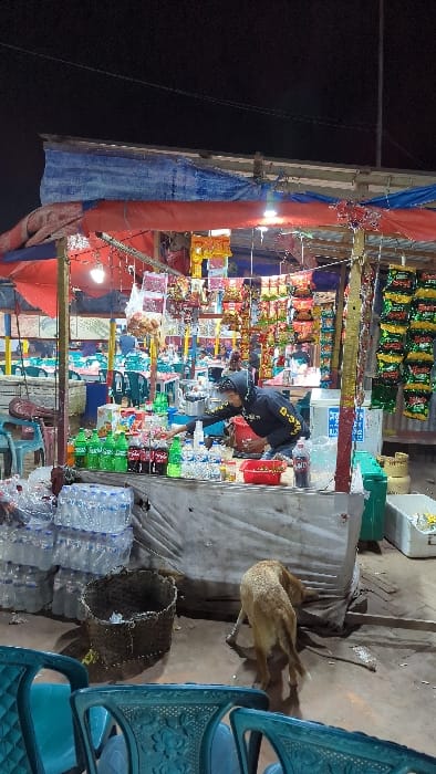 A roadside market stand is stocked with snacks, drinks, and various items. A person wearing a black jacket is near the stand, seemingly shopping or arranging items. To the right, a goat stands near the stand and blue plastic chairs. The stand is lit by overhead lights and appears to be set up at night. Several plastic water bottles are stacked in front of the stand.