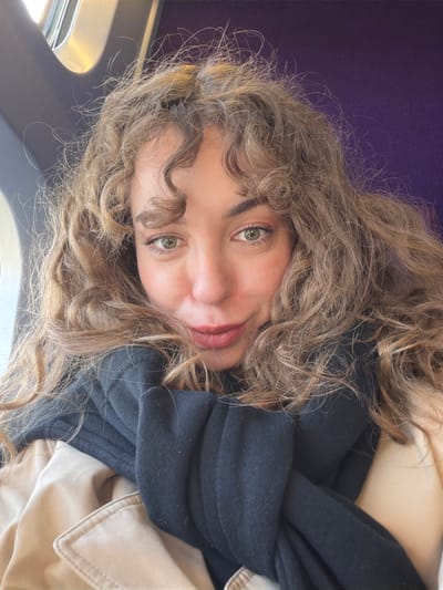A young adult female is captured in a close-up, chest-up portrait, positioned within what appears to be the interior of a vehicle, in Claye-Souilly, France. The subject has fair skin, prominent green eyes, and light brown, voluminous curly hair that frames her face and extends over her shoulders. Her dark eyebrows are well-defined, and her lips are a muted reddish tone, set in a subtle, closed-mouth smile. She is wearing a black fabric garment, likely a scarf or a high-collared top, wrapped around her neck, and a beige or tan coat with visible lapels. She looks directly at the camera. The background is out of focus, showing a curved, light-colored window frame on the left, indicative of a moving conveyance, and a soft, dark purple surface on the right, consistent with vehicle interior paneling or seating.