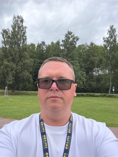 A man with short, grey hair, wearing black-rimmed glasses and a white sweatshirt, stands facing the camera. A blue lanyard with yellow text, reading "Eksjö International Tattoo", is visible around his neck. He is outdoors in a grassy area with trees and a cloudy sky in the background. The location is Aneby, Sweden.