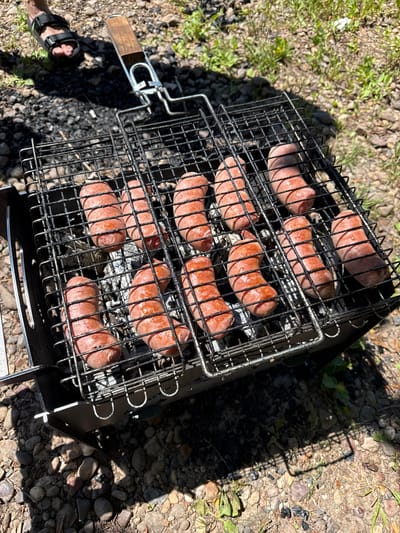 An overhead shot shows hot dogs cooking on a grill over coals. The grill is made of wire mesh and has a wooden handle. The grill is placed on the ground, which is covered with rocks and dirt. In the background, there is some green grass and a white flower. A person's foot wearing a sandal is visible in the upper left corner of the image. The scene appears to be outdoors, possibly in a park or campsite. This image is not specific to any location in the city of Ishimbay, Russia.