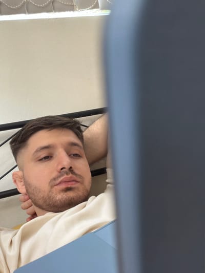 A close-up, upside-down image shows a fair-skinned man with short dark hair and a beard. He is lying down, resting his head on his arm. He is wearing a white top. A dark metal frame runs vertically near his head. Part of a wall is visible to the left, near a string of lights. A blue object occupies a large portion of the top of the frame. No details indicate the location is Pentre, United Kingdom.