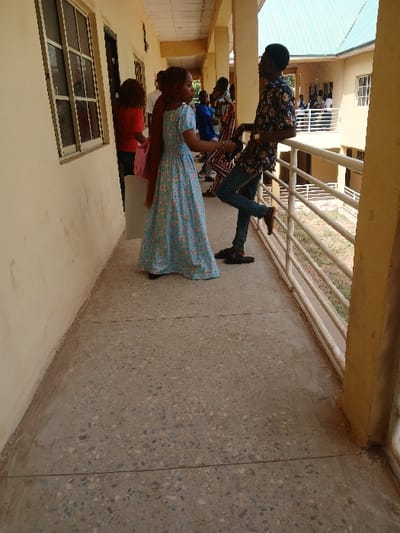 The image depicts a daylight scene on an outdoor covered walkway of a multi-story building in Ugi Majowel, Nigeria.
In the immediate foreground, a female with light brown braids, wearing a light blue long dress with a floral pattern, stands facing a male. The male, with dark hair and skin, wears a dark floral short-sleeved shirt, blue jeans, and dark slide sandals; he is leaning against a white metal railing with his left elbow, his right foot resting on a lower bar of the railing. Both individuals appear to be engaged in conversation.
The walkway features a light beige painted wall on the left, which contains a large multi-paned window and a dark-colored door. The floor is light-colored concrete with visible aggregate. The white metal railing on the right side of the walkway borders an open courtyard area.
In the background, further down the corridor, several other individuals are present. A female in a red shirt is partially visible near a doorway on the left, carrying a light pink bag. Other figures are indistinctly visible standing or leaning against railings. Across the courtyard, a separate building with a light green corrugated metal roof and white walls is visible, with additional individuals on its lower balcony.
