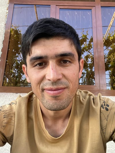 A man with dark hair and brown eyes is centered in the image, looking directly at the camera. He has a light complexion and some facial hair. He is wearing a light brown, crew-neck sweatshirt with an anime-style design on the sleeve. Behind him is a window with a brown frame. Reflections of trees and foliage are visible in the glass panes. The background context implies an outdoor or partially outdoor setting. There is no other explicit information visible indicating that it is taken in Akhtachi, Uzbekistan, or indicating there is an action or interaction taking place.