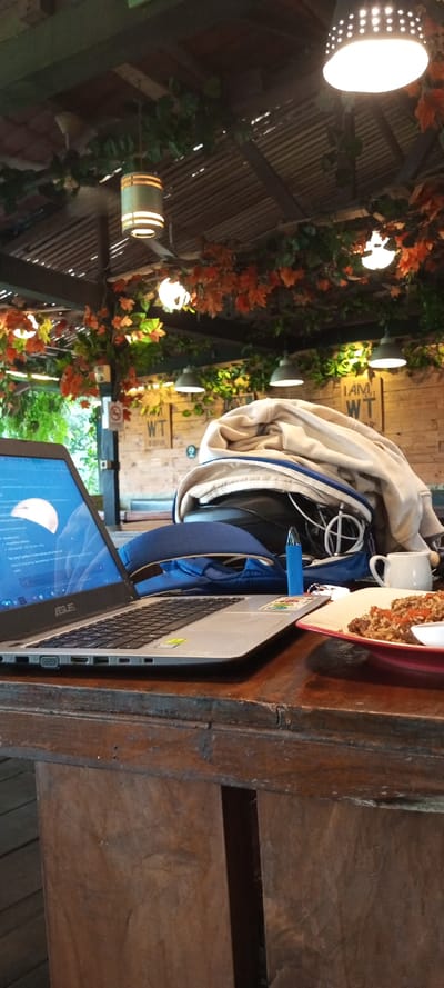 An ASUS laptop sits open on a dark wooden table, next to a plate of food and a small white cup. A blue backpack with a tan jacket is piled behind it. The background shows a wooden structure with plants and decorative lighting overhead. Some writing, "I AM WT," is visible on a wooden wall in the background.