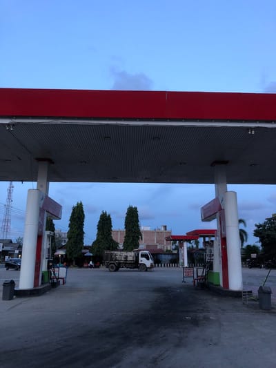 The image depicts an exterior view of a Pertamina gas station in the city of Matang, Indonesia. The scene is captured at dusk or early morning under a cloudy sky. In the foreground, the asphalt ground is visible, with tire marks and some debris. A gas pump structure with a red canopy and white support pillars is prominently featured. One of the pumps has a red and white sign.
To the left of the gas pump, a black trash can is placed on the ground. In the middle ground, several tall, slender trees stand in a row. Behind the trees, a white truck with a rusted cargo bed is parked. Further in the background, buildings with light-colored walls are visible, along with a telecommunications tower on the far left. Scattered tables and chairs, some with umbrellas, suggest an outdoor seating area. People are present in the background, appearing as small figures sitting at tables or walking.
To the right of the central gas pump, another similar pump structure is visible. A red sign with white text is mounted on a post. A metal rack with a green object is next to the pump. Further to the right, a trash can and a motorcycle are discernible. A sign with "TUTUP SEBENTAR" and "JUM'AT" is positioned near the central area. The overall environment suggests a typical urban setting with commercial activity.