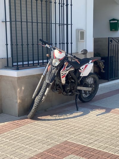 A dirt bike is parked on a sidewalk in front of a building. The building has a white facade and a black metal gate over a window. To the right of the gate, a green mailbox is mounted on the wall. The sidewalk is paved with rectangular bricks in shades of beige and reddish-brown. The dirt bike is black, white, and red, with knobby tires. The motorcycle is leaning on its kickstand. The scene appears to be in a residential area.