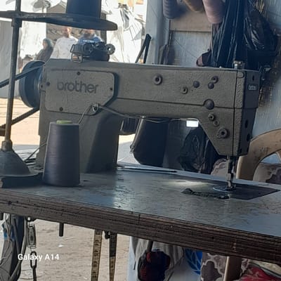 The image shows a Brother brand sewing machine resting on a wooden table. A spool of thread is attached to the machine. The scene appears to be outdoors, potentially in Al-Mawasi, although specific location details are unidentifiable. Additional items are present in the background, including fabric and clothing. A measuring tape is seen near the edge of the table. The image also includes a watermark indicating it was taken with a Galaxy A14.