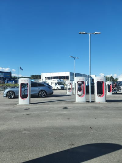 The image depicts a row of Tesla charging stations situated in an outdoor parking lot. The charging stations are white with red accents and prominently display the Tesla logo. Several electric vehicles are parked at the stations, suggesting they are actively being charged. In the background, there are buildings and trees, indicating an urban or suburban setting. A clear blue sky suggests fair weather and daytime. The presence of streetlights and the overall arrangement of the charging infrastructure point towards a dedicated electric vehicle charging hub. The scene is set in Strömstad, Sweden. No people are visible in the image, and the focus is on the charging infrastructure and the vehicles.