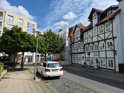 The image depicts a street scene in Brunswick, Germany, specifically showing a section of the city that features both modern and historic architecture. On the left side of the street, a modern apartment building with a pale yellow and blue facade stands tall. In front of this building, several large green trees provide shade. Adjacent to the modern building is a street with a paved sidewalk.
The right side of the street showcases traditional German half-timbered houses with white plaster walls and dark wooden beams. These houses appear to be occupied by businesses, as indicated by signage visible on some of the buildings. One building displays a sign that reads "Praxis für Medicinische Kosmetik" (Practice for Medical Cosmetics).
A white station wagon is parked on the side of the road. A bicycle is leaning against the facade of one of the half-timbered houses. Further down the street, scaffolding and white sheeting suggest that a building is undergoing renovation or construction. The sky is blue with scattered white clouds, indicating a pleasant day. The street itself is paved with cobblestones in some areas and asphalt in others. Traffic signs and street lamps are also present.