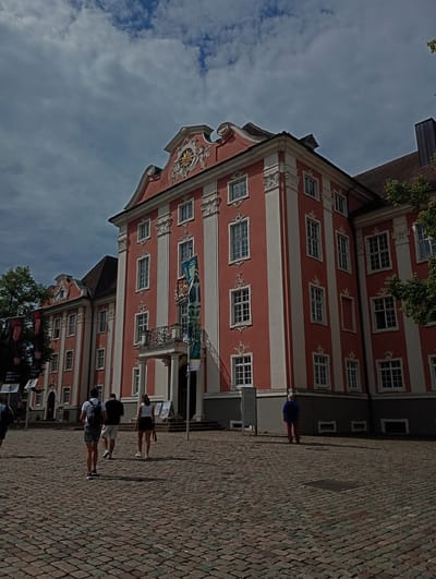 The image shows the southern facade of the Neues Schloss (New Castle) in Meersburg, Germany. The building is painted in a salmon pink with white vertical stripes and ornate white architectural details around the windows and cornices. A prominent gold clock is visible on the central gable. The building has multiple stories with numerous windows. A balcony with an iron railing extends from the central section of the building. Several banners hang from the facade, one displaying blue and white graphics and another with a portrait. A cobblestone plaza is in the foreground, with three individuals walking away from the camera across it. One person on the left wears a white shirt, black shorts, and a backpack. The middle person wears a black t-shirt and white shorts. The person on the right wears a white top and dark shorts. A fourth person, viewed from the back, stands further to the right in the plaza, wearing a blue shirt and red pants. The sky above is cloudy with patches of blue. Trees are visible on the far right and left edges of the image.